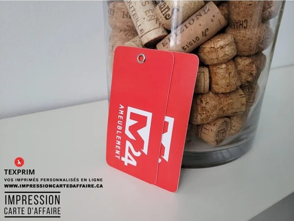 Deux étiquettes cartonnées rouges à coins arrondis et œillet métallique, devant un contenant en verre.
